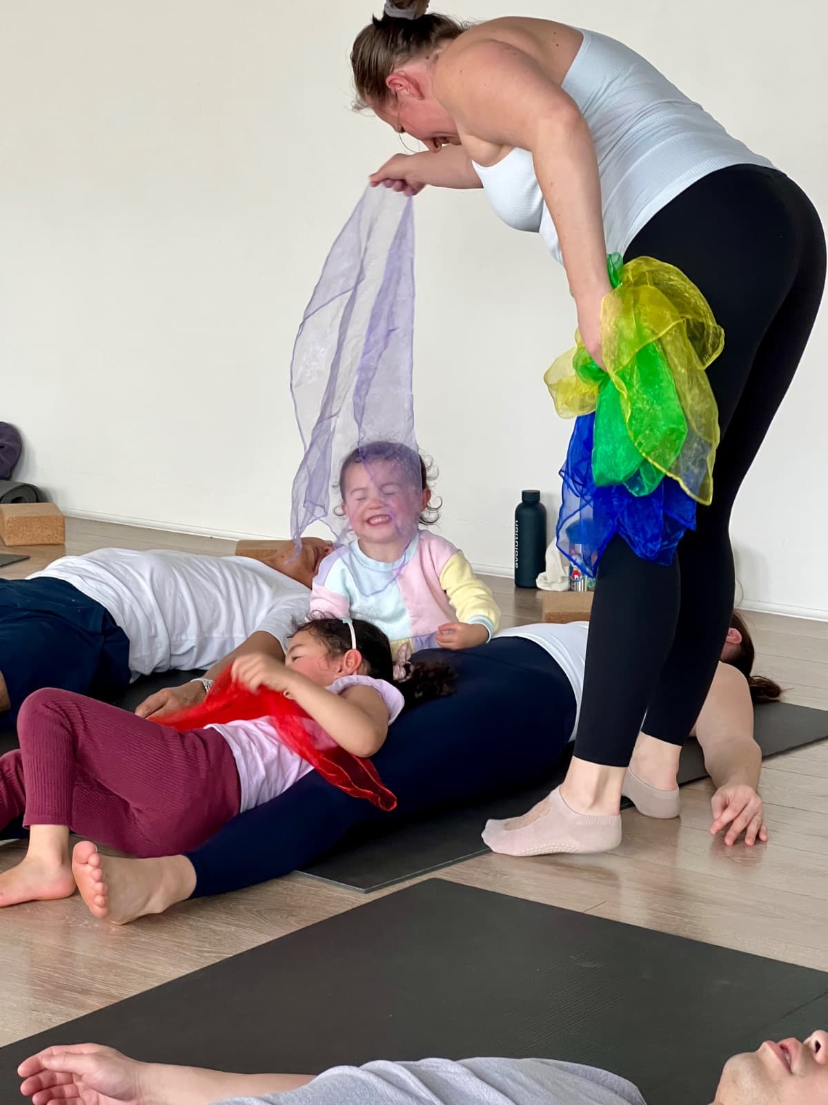 Teacher playing with colourful scarves as children giggle on the mat